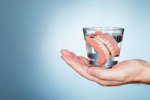 hand holding a glass of water containing dentures in front of a light blue background